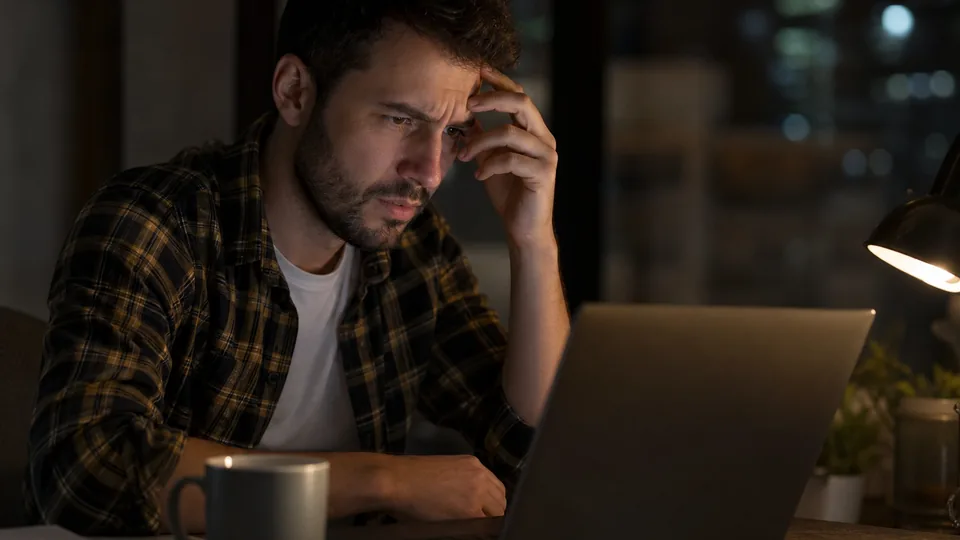 Adult man at a desk at night looking puzzled at a laptop that will not load a site