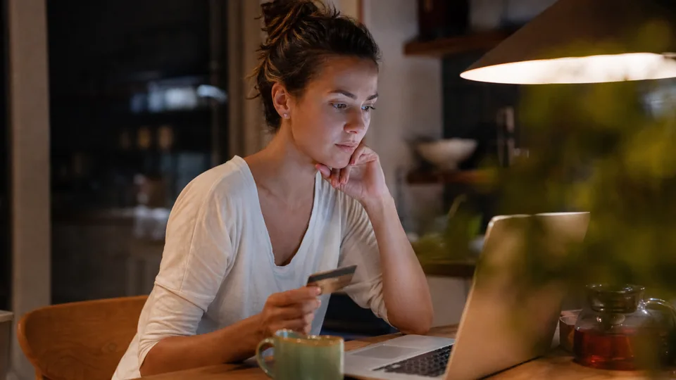 Adult woman at a kitchen table at night with a laptop and a payment card, checking a subscription charge