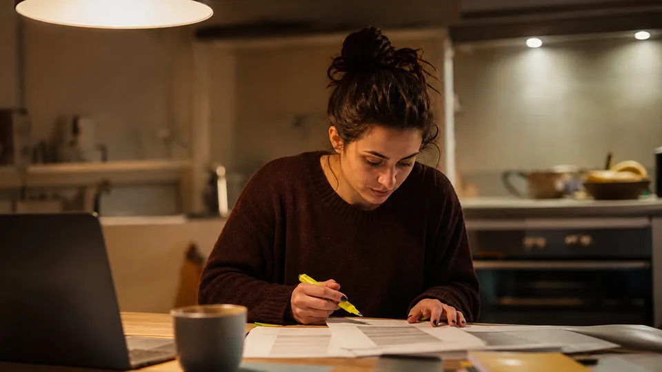 Adult woman at a kitchen counter at night comparing two printed pages with a highlighter